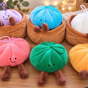 May include: Six plush dumpling toys with smiling faces, each featuring a small loop on top and two brown legs. Three are nestled in brown fuzzy baskets, while the pink, green, and yellow ones sit separately. Each toy has a soft, rounded shape.