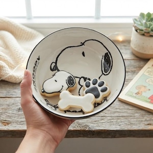 Puede incluir: Un cuenco redondo de cerámica blanca con un dibujo de líneas negras del personaje Snoopy y el texto SNOOPY impreso en el lateral. Dentro del cuenco hay tres galletas de azúcar con forma de cabeza de perro, huella y hueso con glaseado blanco.