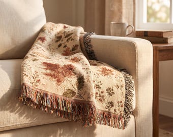 May include: A woven throw blanket featuring a cream-colored base with a detailed pattern of autumn leaves in shades of rust, orange, and brown. The blanket has a textured appearance and is finished with multi-colored fringe along the edges.