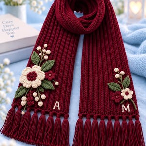 May include: A deep burgundy ribbed knit scarf featuring crocheted floral embellishments in cream, dark red, and green. The scarf ends are finished with long matching tassels. The letters A and M are embroidered in white on the lower sections.