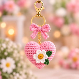 May include: A pink crocheted heart keyring featuring a small pink crocheted bow at the top and a white daisy flower with a yellow centre and two green leaves attached to the front, all suspended from a gold-toned metal lobster clasp.