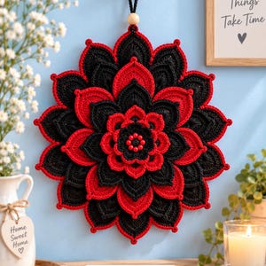 Può includere: Una decorazione da parete circolare all'uncinetto con un motivo floreale a più strati di petali in filato nero e rosso alternati. Il centro è accentuato da piccole perline rosse e l'oggetto è appeso a un cordino nero su un gancio in legno.