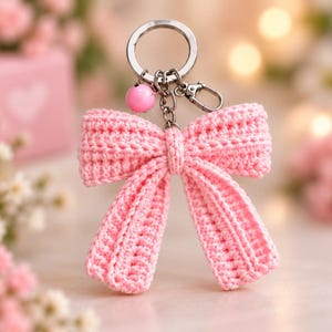 May include: A light pink crocheted bow keychain featuring a silver-toned metal split ring, a small lobster clasp, and a single round pink bead charm attached to the chain. The bow is constructed with a textured yarn stitch pattern.