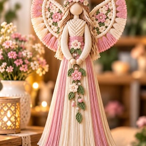 May include: A macrame angel ornament featuring cream and pink cord. The figure has a floral crown, intricate wing patterns with pink accents, and a long dress with vertical pink stripes. A floral garland with green leaves hangs from the hands.