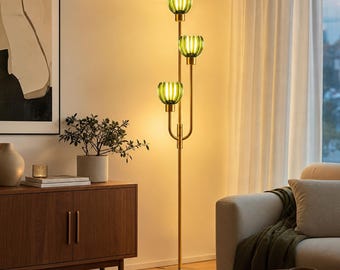 May include: A tall floor lamp with a slender gold-toned metal stand and three vertical arms. Each arm holds a bowl-shaped, ribbed green glass shade that emits a warm, glowing light. The lamp features a circular base and a vertical design.