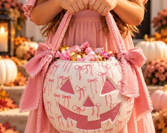 May include: A round white pumpkin-shaped bucket featuring a pink bow pattern, a carved jack-o'-lantern face with pink cutouts, and two large pink fabric bows attached to the sides. The bucket is filled with various gold and pink wrapped sweets.
