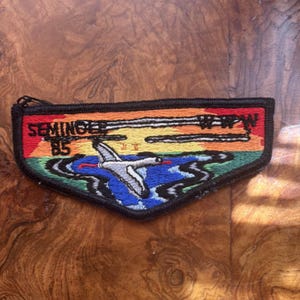 May include: A triangular embroidered patch featuring a sunset landscape with a flying white bird over blue water. The text SEMINGEE 85 and WPW is stitched in black thread. The patch has a thick black border and a zipper pull at the top left corner.