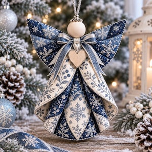 May include: A fabric angel ornament featuring a quilted navy blue and white snowflake pattern. The angel has a round wooden bead head, a central wooden heart accent, and a decorative bow at the neck. It hangs from a silver cord loop.