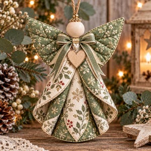 May include: A hanging angel ornament crafted from quilted fabric featuring a green and cream botanical pattern with snowflakes and red berries. The figure includes a smooth wooden bead head, a fabric bow at the neck, and a small wooden heart charm.