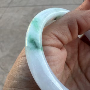 May include: A smooth, circular bangle bracelet crafted from polished stone. The bangle features a translucent white base color with distinct, vibrant green patches and streaks distributed throughout the material, creating a natural marbled appearance.