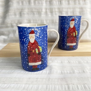 May include: Two cylindrical ceramic mugs featuring a deep blue background with white painted snowflakes. Each mug displays a central illustration of Santa Claus wearing a red coat and holding a green gift box, complete with a white handle.