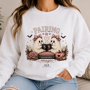 May include: A white long-sleeve sweatshirt featuring a graphic of two ghosts with pink bows holding mugs. The text reads "PAIRING is magic" and "PAIRING REINFORCEMENT" with "BS BEHAVIOR SOCIETY" at the bottom. The design includes candles, roses, and pumpkins.