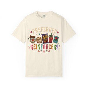 Op de afbeelding: Een crèmekleurig t-shirt met een kleurrijke grafische print. Het ontwerp bevat een mok, een concha-broodje, een zak Takis, een drankje en een rozenkaart, met de tekst PREFERRED REINFORCERS in een meerkleurig lettertype en kleine hartjes.