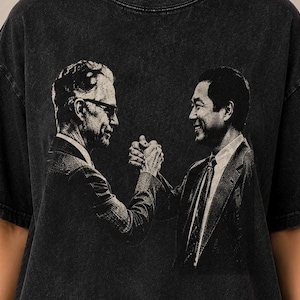 May include: A black, acid-washed short-sleeve t-shirt featuring a monochromatic graphic print of two men in suits shaking hands. The design is rendered in a high-contrast, grainy black and white style, centered on the front of the garment.