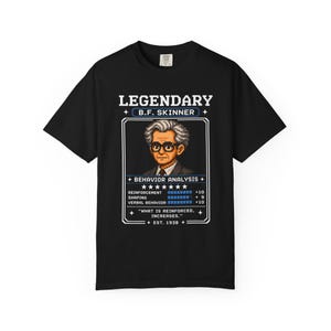 Op de afbeelding: Een zwart t-shirt met een pixel art afbeelding van B.F. Skinner. Het ontwerp bevat de tekst LEGENDARY B.F. SKINNER, BEHAVIOR ANALYSIS, statistieken voor reinforcement, shaping, verbal behavior en het citaat WHAT IS REINFORCED, INCREASES.