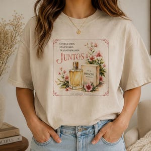 May include: A beige short-sleeve t-shirt featuring a graphic print of a perfume bottle and a box labeled IMPACTO, surrounded by pink flowers. The text reads CONECTAMOS, ENSEÑAMOS, TRANSFORMAMOS, JUNTOS, and THE BEHAVIOR SOCIETY in a decorative frame.