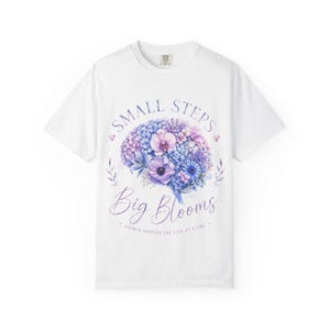 Op de afbeelding: Een wit t-shirt met korte mouwen en een centrale afbeelding van een menselijk brein samengesteld uit blauwe, paarse en roze bloemen. De tekst luidt "SMALL STEPS Big Blooms" en "GROWTH HAPPENS ONE STEP AT A TIME" in een paars script- en schreeflettertype.