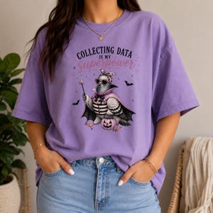 Può includere: Una t-shirt a maniche corte color viola chiaro con la grafica di un piccione che indossa occhiali da sole, un fiocco e un mantello, che tiene in mano una bacchetta a stella e un secchiello a forma di zucca. Il testo recita "COLLECTING DATA IS MY SUPERPOWER".