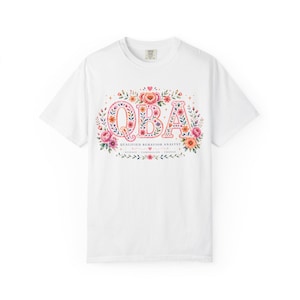 Op de afbeelding: Een wit t-shirt met korte mouwen voorzien van een bloemmotief met de letters QBA in een roze en oranje patroon. Onder de letters staat de tekst QUALIFIED BEHAVIOR ANALYST, gevolgd door de woorden SCIENCE, COMPASSION, CHANGE met kleine hartjes.
