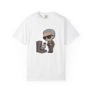 Op de afbeelding: Een wit t-shirt met korte mouwen voorzien van een grafische illustratie van een gestileerd figuur in een bruin pak en zwarte zonnebril met een notitieblok, naast een grijze machine met rode knoppen en een duif met een zonnebril.
