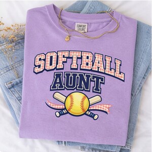 May include: A lavender t-shirt featuring the text "SOFTBALL AUNT" in a navy blue and pink gingham font. Below the text is a graphic of a yellow softball with red stitching resting in front of two crossed bats and a pink gingham ribbon.