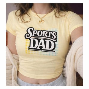 May include: A pale yellow short-sleeved t-shirt featuring the text SPORTS DAD in bold black letters. The text is layered over a light yellow and blue checkered square pattern. The shirt has a crew neckline and a fitted silhouette.