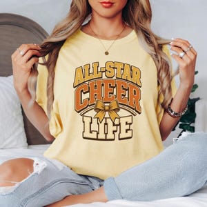 Op de afbeelding: Een lichtgeel t-shirt met korte mouwen en de tekst ALL-STAR CHEER LIFE in dikke oranje en crèmekleurige blokletters. Een afbeelding van een goudgele strik staat in het midden tussen de woorden CHEER en LIFE op de voorzijde.