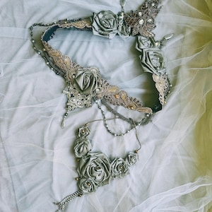 May include: A decorative accessory set featuring a wide band and a smaller piece, both adorned with sage green fabric roses, intricate gold-toned lace trim, metallic beadwork, and dangling crystal-like accents connected by thin silver-toned chains.