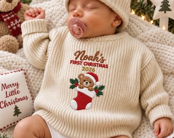 May include: A cream-colored ribbed knit sweater featuring an embroidered design of a teddy bear in a red Christmas stocking with the text Noah's FIRST CHRISTMAS 2026. The sweater is worn with a matching cream-colored knit beanie hat.