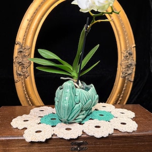 Peut inclure: Un cache-pot en céramique vert clair brillant en forme de grenouille contenant une orchidée blanche aux feuilles vertes. Le cache-pot repose sur un napperon au crochet à motifs circulaires beiges et bleu sarcelle sur une boîte en bois.