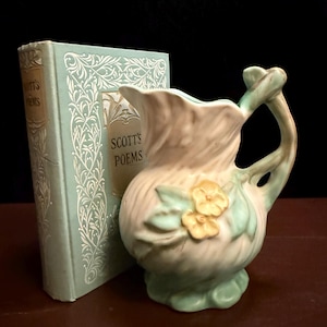 May include: A light green and cream ceramic pitcher with a molded floral design featuring two yellow flowers and a curved handle, positioned next to a light green hardcover book with the text "SCOTT'S POEMS" printed on the front cover.