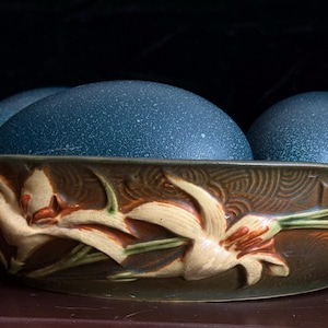 Puede incluir: Un cuenco de cerámica con esmalte marrón y relieves florales en crema y verde. El cuenco contiene tres huevos grandes de color azul oscuro con motas. El exterior del cuenco presenta intrincados patrones de remolinos tras el diseño floral.