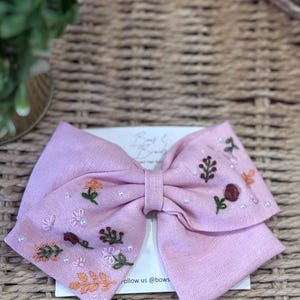 May include: A light pink linen hair bow featuring embroidered floral motifs in shades of orange, green, and dark red, accented with small white beads. The bow is attached to a white display card with the text "Bows & Beads" and "Follow us @bows".