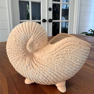 May include: A pale peach-colored ceramic vase shaped like a nautilus shell. The exterior features a textured, granular surface with raised ridges and a spiral design. The vessel stands on three small, sturdy feet and has an open top cavity.