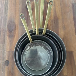 Pode incluir: Um conjunto de quatro frigideiras redondas de metal encaixadas, cada uma com um cabo longo e fino em tom de latão com um orifício em forma de gota na extremidade. As frigideiras têm um acabamento metálico escuro no exterior e um interior prateado liso.