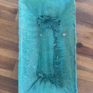 Könnte beinhalten: Ein rechteckiges Keramiktablett mit einer leuchtend türkisfarbenen Glasur und dunkelblauen Akzenten. Die Oberfläche weist ein strukturiertes, gesprenkeltes Finish auf, bei dem sich dunklere Pigmente in der Mitte sammeln und zu den Rändern hin auslaufen.