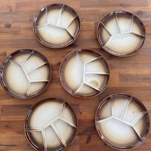 May include: A set of six round ceramic divided plates featuring a gradient design that transitions from a dark brown rim to a light cream center. Each plate is partitioned into four distinct sections by raised dividers, creating a functional layout.