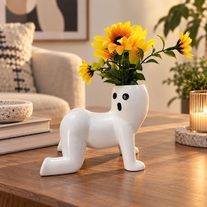 May include: A white ceramic vase shaped like a four-legged creature in a crawling position with a surprised facial expression featuring two black eyes and an open mouth, filled with a bouquet of bright yellow sunflowers with green stems and leaves.