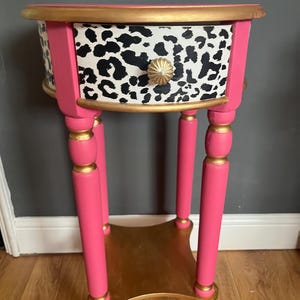 May include: A round side table featuring a bright pink frame with gold-painted accents on the legs and trim. The single drawer front displays a black and white leopard print pattern, complemented by a round, fluted gold-coloured metal drawer knob.