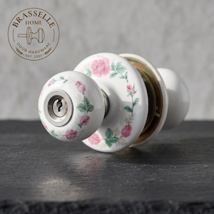 May include: A white porcelain door knob set featuring a delicate pink rose and green leaf floral pattern. The front knob includes a central silver-toned keyhole mechanism. The logo BRASSELLE HOME DOOR HARDWARE EST. 2023 is visible in the upper left corner.
