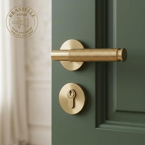 May include: A brushed brass door handle and matching keyhole escutcheon mounted on a dark green door. The handle features a cylindrical grip with a horizontal ribbed texture. The Brasselle Home Door Hardware logo is visible in the top corner.