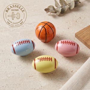 Könnte beinhalten: Vier Keramik-Möbelknöpfe mit Sportmotiven. Ein runder, orangefarbener Basketball-Knopf und drei ovale, footballförmige Knöpfe in Hellblau, Rosa und Gelb, jeweils mit roten Schnürsenkeln. Das Logo BRASSELLE HOME DOOR HARDWARE EST. 2023 ist zu sehen.