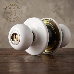 May include: A white ceramic door knob set featuring a round handle with a central brass keyhole mechanism. The assembly includes a circular white backplate and a metallic internal mounting plate with a gold-toned finish, visible from the side.