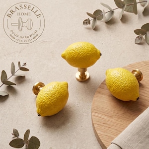 May include: Three lemon-shaped cabinet knobs with textured yellow surfaces and gold-toned metal bases. The knobs are arranged on a light surface, with a logo in the top left corner reading BRASSELLE HOME DOOR HARDWARE EST. 2023.