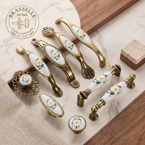 Op de afbeelding: Een collectie kastbeslag van antiek messing met witte keramische inzetstukken met gele bloemmotieven. De set bevat diverse ladehandgrepen, grepen en knoppen met gedetailleerde metalen afwerkingen en het Brasselle Home logo.