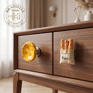 May include: Two decorative cabinet knobs mounted on a wooden drawer. One knob is a yellow, circular egg tart shape. The other knob is a beige fabric-textured bag containing three miniature baguettes with the word Pain printed on the front.