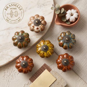 May include: Six round, pumpkin-shaped ceramic cabinet knobs with a glossy, multi-colored glaze finish and a central bronze-toned metal cap. The knobs feature varied colors including brown, yellow, green, and blue, arranged on a light surface.