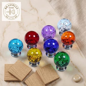 May include: A collection of eight spherical glass cabinet knobs in various colors including red, blue, yellow, green, purple, and clear, each featuring internal air bubbles and a polished silver-tone metal base, arranged on a marble surface.