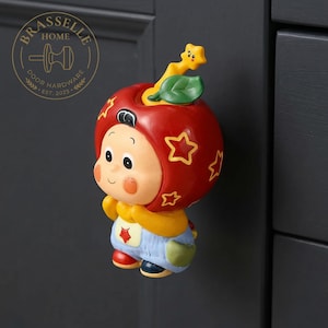 May include: A decorative cabinet knob shaped like a cartoon character wearing a red apple-shaped hat with yellow star outlines. The figure has a yellow body, light blue overalls with a red star, and a small green leaf and yellow star on top.