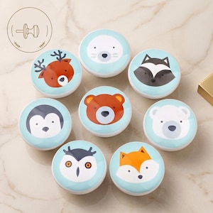 May include: A set of eight round cabinet knobs featuring colorful illustrations of woodland animals including a deer, bear, raccoon, penguin, polar bear, owl, and fox on a light blue background. Each knob has a smooth, glossy finish.